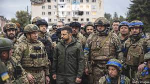 A man in a dark jacket stands among a group of soldiers in camouflage uniforms and helmets, looking serious. The overlaid…
