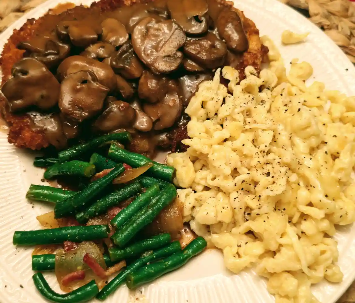 A close-up photo of a white plate holding a meal of breaded meat covered in mushroom gravy, creamy pasta, and sautéed green…