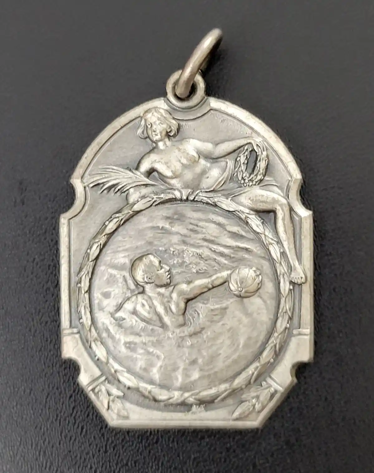 A silver medal features a classical nude woman holding a laurel wreath above a circular scene of a man kicking a ball…