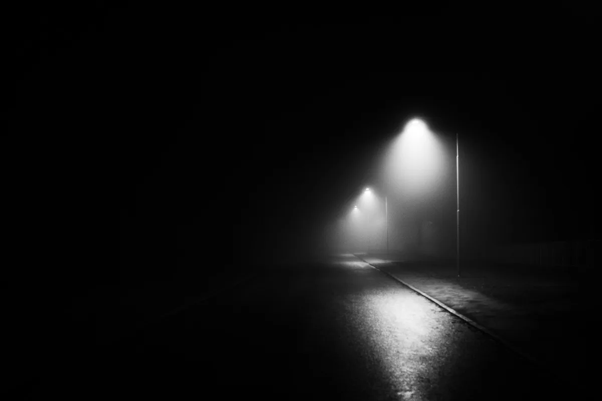 A black and white photo of a foggy, empty street at night, illuminated by a few distant streetlights casting beams into the…