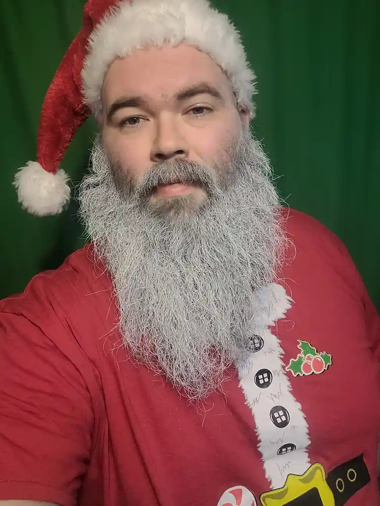 A man with a long, gray beard and mustache wears a red Santa hat and a red t-shirt with a Santa Claus design, including a…