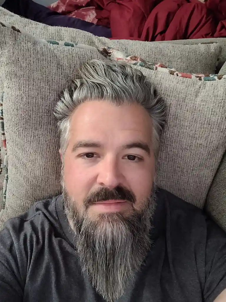 A man with gray hair and beard lies on a textured gray couch, looking directly at the camera with a neutral expression…