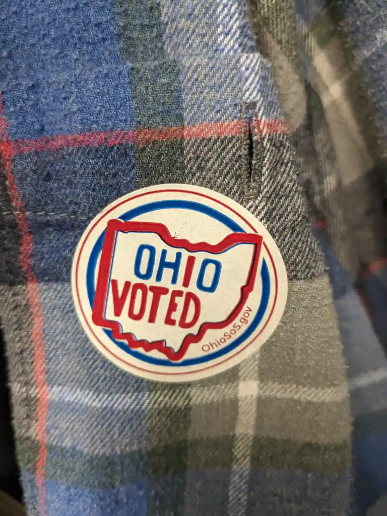 A circular 'OHIO VOTED' sticker with red and blue borders and the outline of the state of Ohio is stuck on a blue, gray, and…
