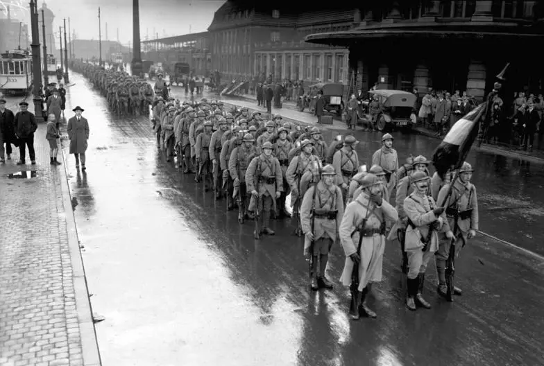a black and white historical photo shows a long column of soldiers marching down a wet city street, with civilians watching…