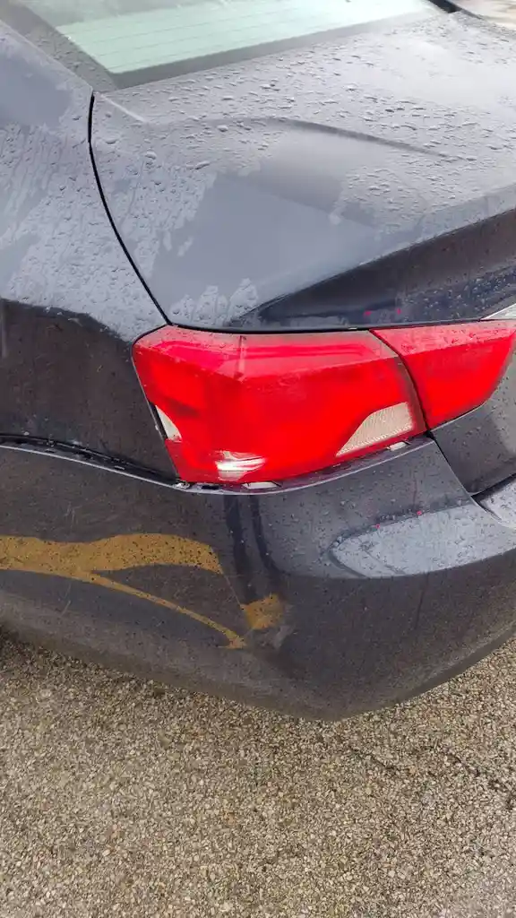 A close-up of the rear of a dark gray car covered in rain droplets, showing a damaged red taillight and a yellow…