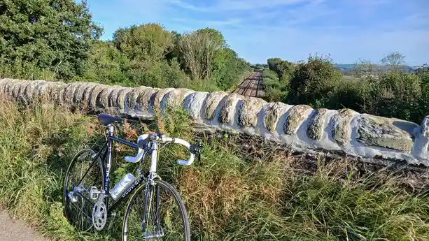 A black and white road bicycle is parked on grass beside a stone railway bridge, with train tracks receding into the…