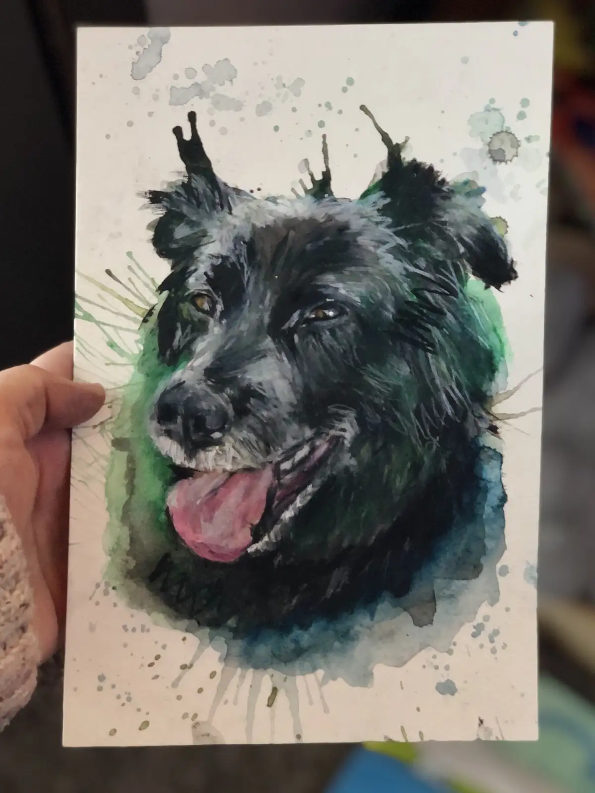 A hand holds up a watercolor painting of a black dog with a happy, panting expression, surrounded by green and blue paint…