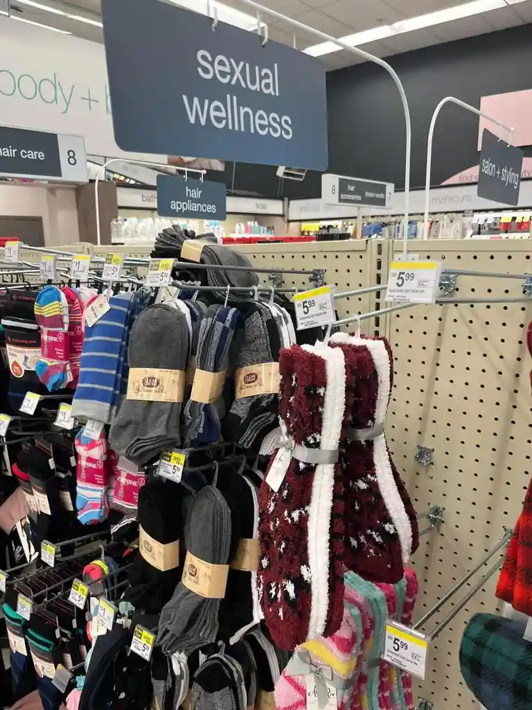 A store aisle displays various socks on racks under a large blue sign that reads 'sexual wellness', with other signs for…