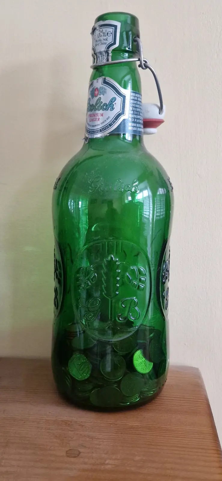 A green glass bottle of Coors Premium Lager with a swing-top cap sits on a wooden surface, filled with numerous coins…