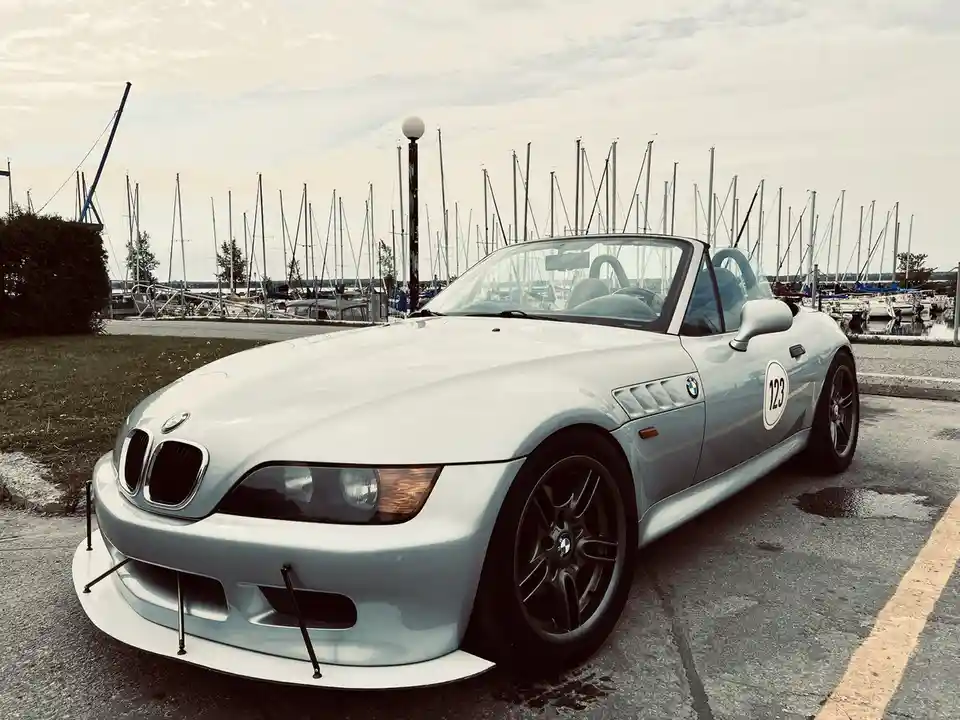 A light blue BMW Z3 roadster with racing number 123 on its side is parked in a lot with a marina full of sailboats visible…
