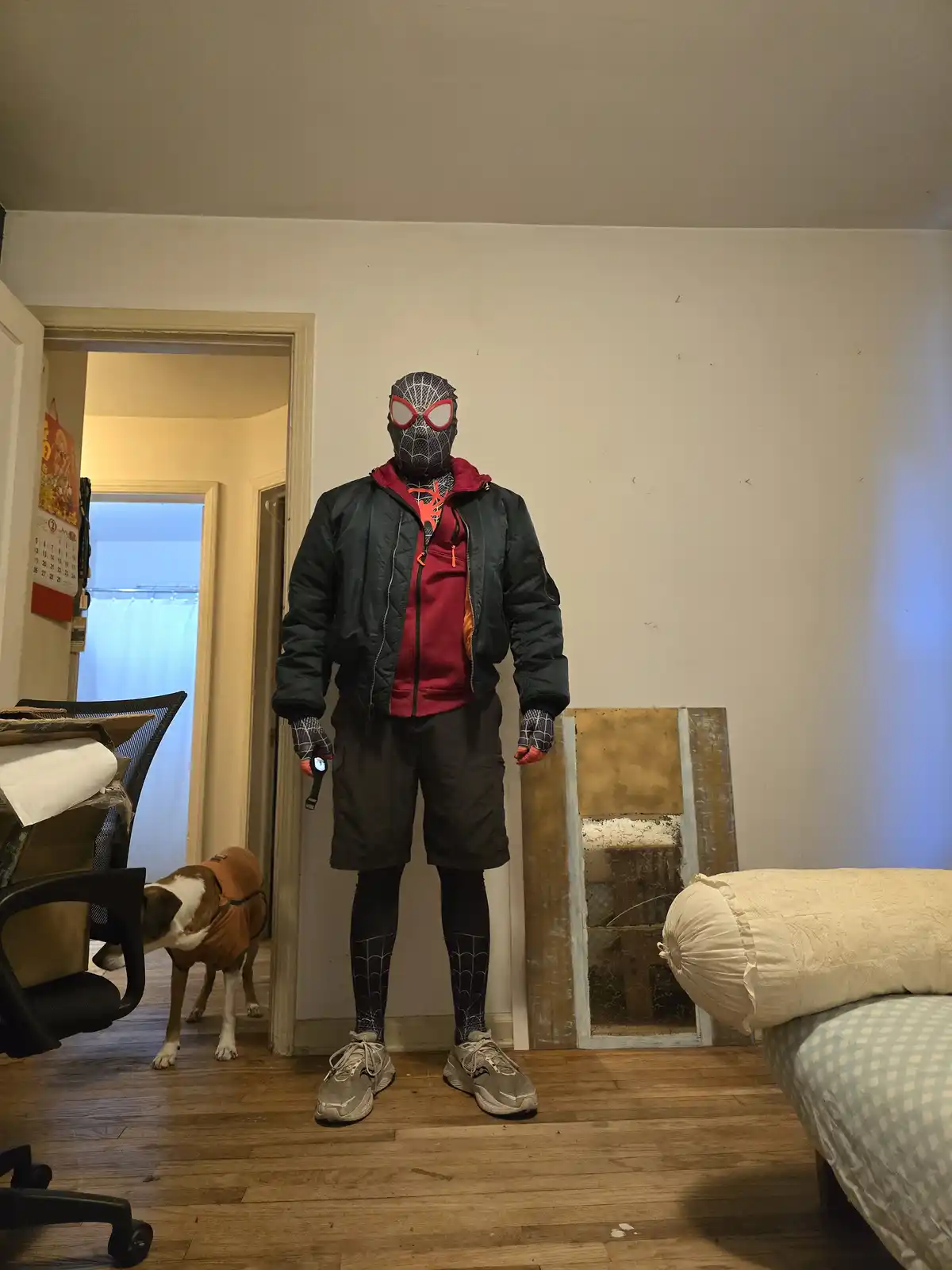A person wearing a Spider-Man mask, red hoodie, black jacket, shorts, and leggings stands in a bedroom. They hold a…