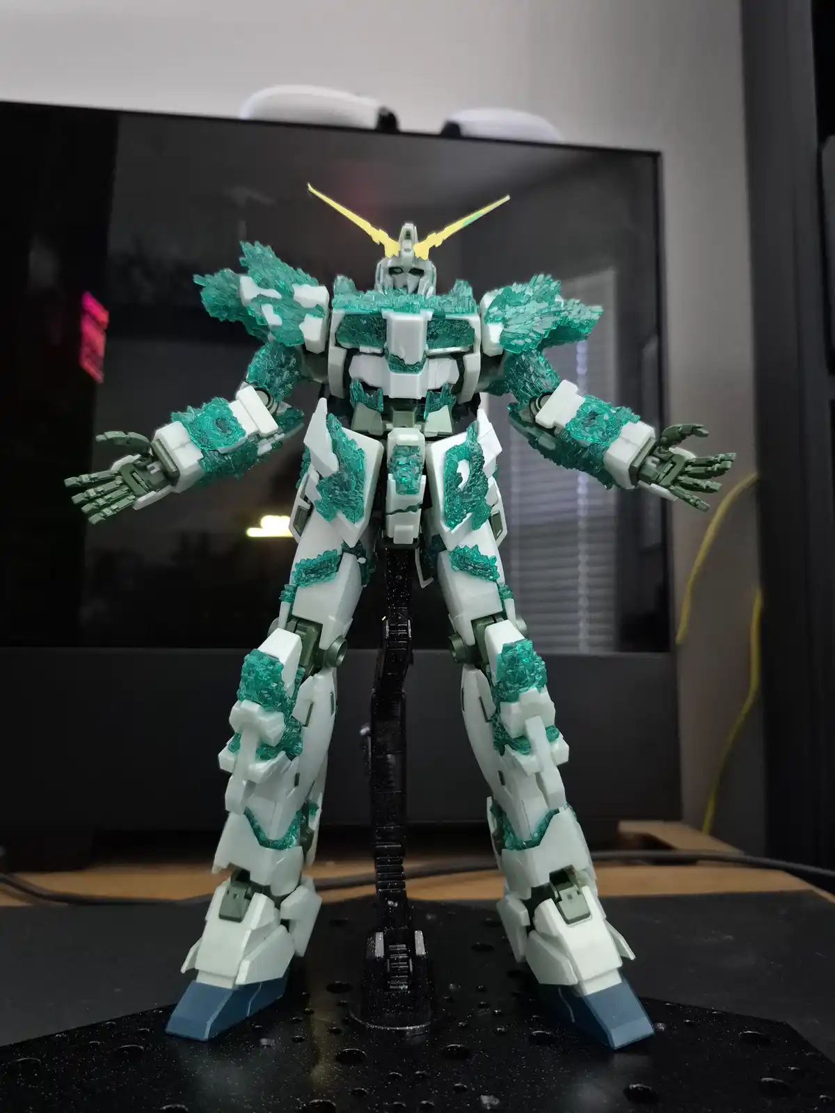 A white and teal Gundam model with crystalline green armor pieces is posed on a black display stand in front of a dark…