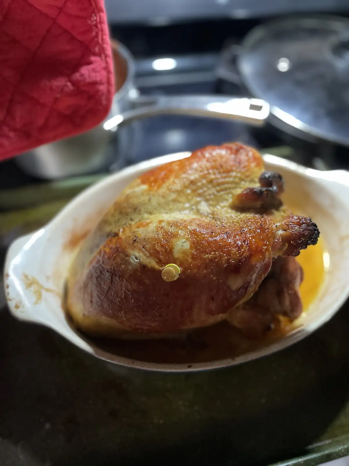 A roasted chicken sits in a white ceramic baking dish, its golden-brown skin glistening with juices. A small yellow button…