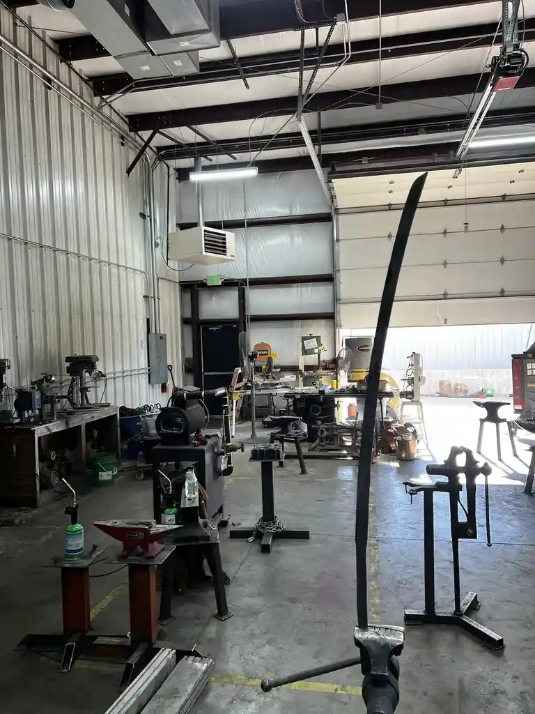 A wide-angle photo of a metal workshop filled with tools, anvils, and machinery, with a long, crudely shaped sword-like…