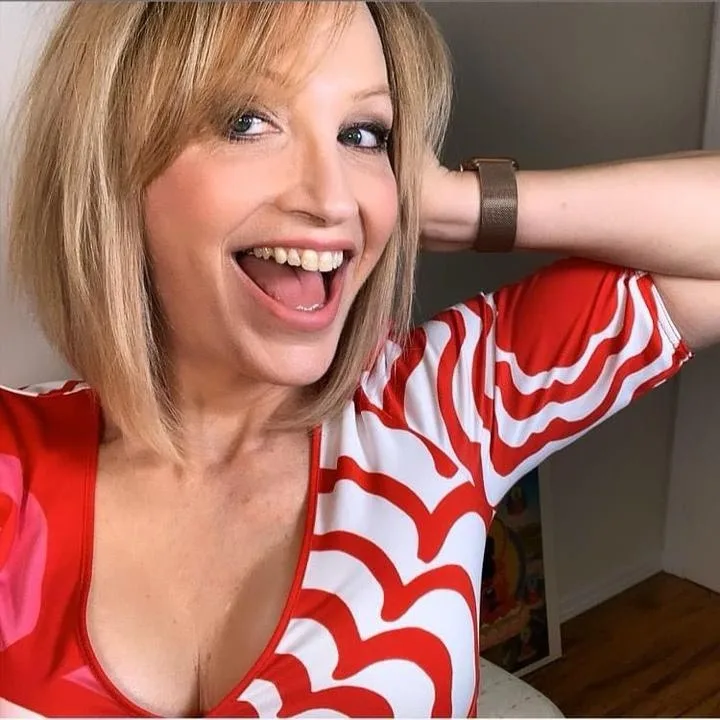 a woman with blonde hair and a red-and-white patterned top is smiling widely with her mouth open, one arm raised behind her…
