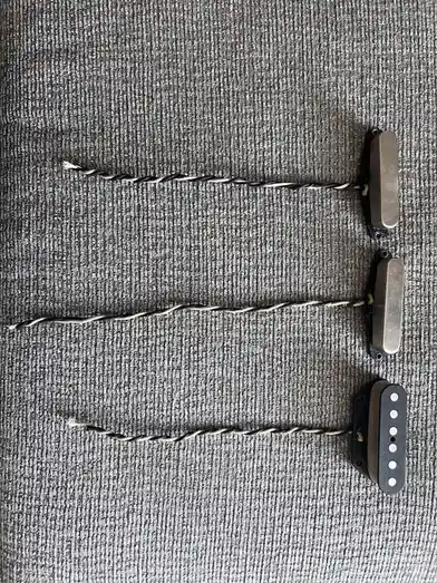 Three electric guitar pickups are laid out vertically on a gray textured fabric, each connected by a twisted black-and-white…