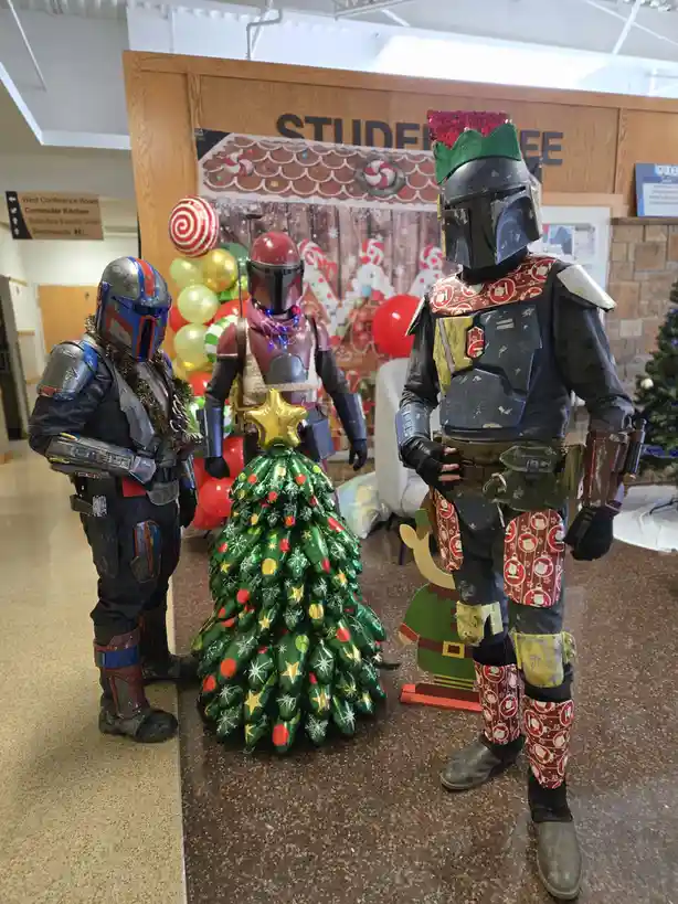 Three people in Mandalorian armor stand around a green foil Christmas tree decorated with stars and snowflakes. The person…