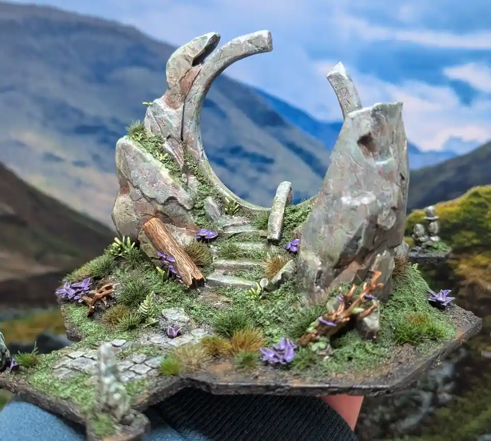 A hand holds a detailed miniature diorama featuring mossy terrain, purple flowers, small stone ruins, and a large, broken…