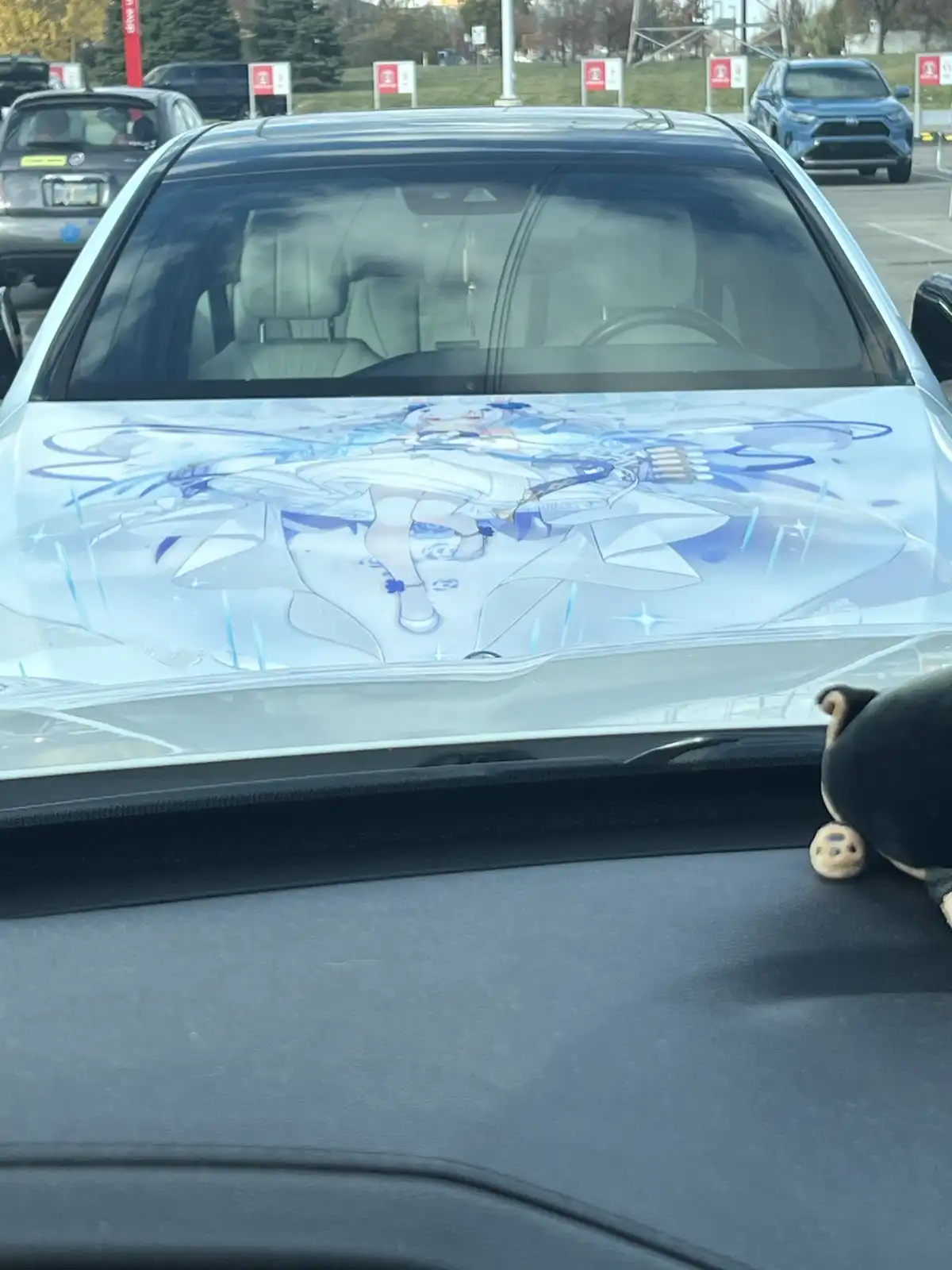 A silver car's hood is covered in a large, detailed anime-style illustration of a blue-haired female character in a white…
