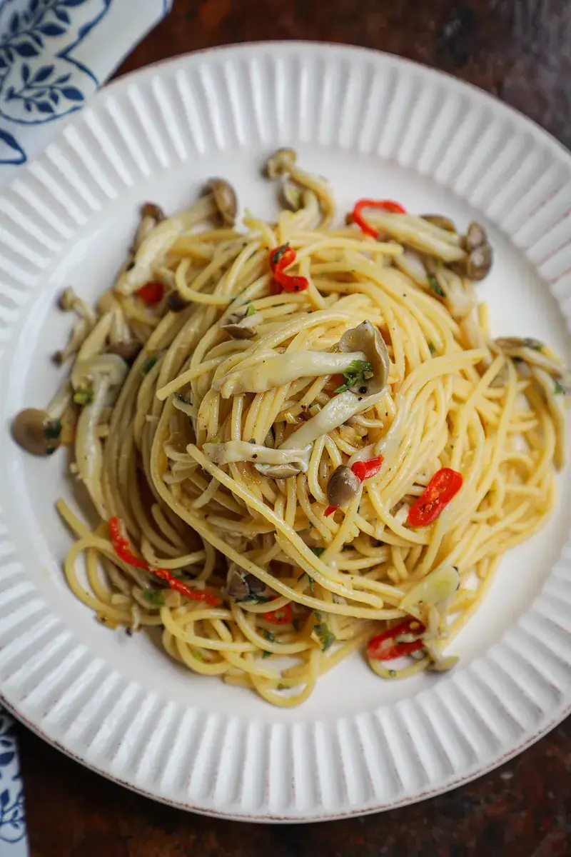 A close-up photo of a white plate with pasta, mushrooms, and red chili peppers. The pasta is mixed with sliced mushrooms and…