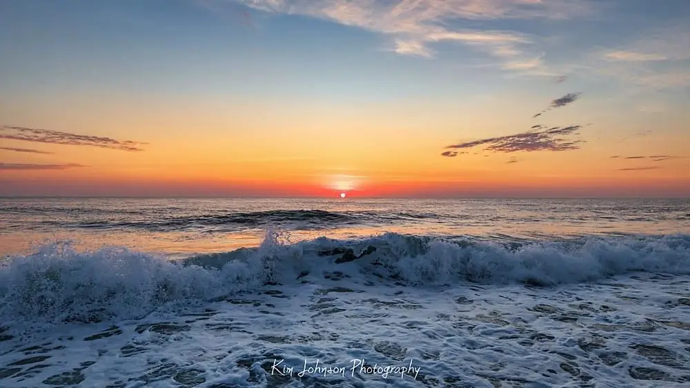 A serene sunset over the ocean with waves crashing in the foreground, the sun low on the horizon casting orange and blue…