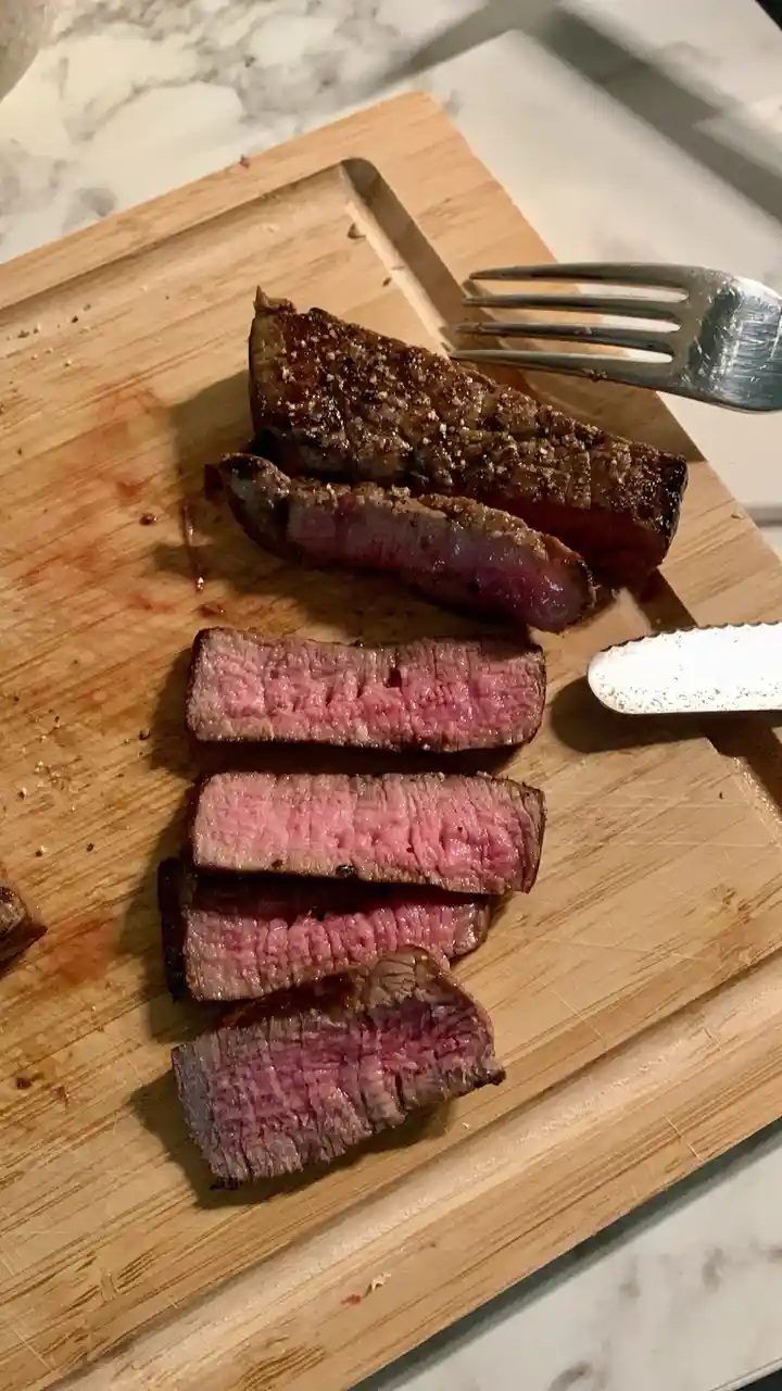 A close-up photo shows sliced medium-rare steak on a wooden cutting board, with a fork and knife nearby, revealing…