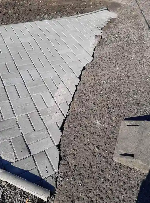 A close-up photo shows a sharply angled crack dividing a paved sidewalk made of grey interlocking concrete pavers from an…