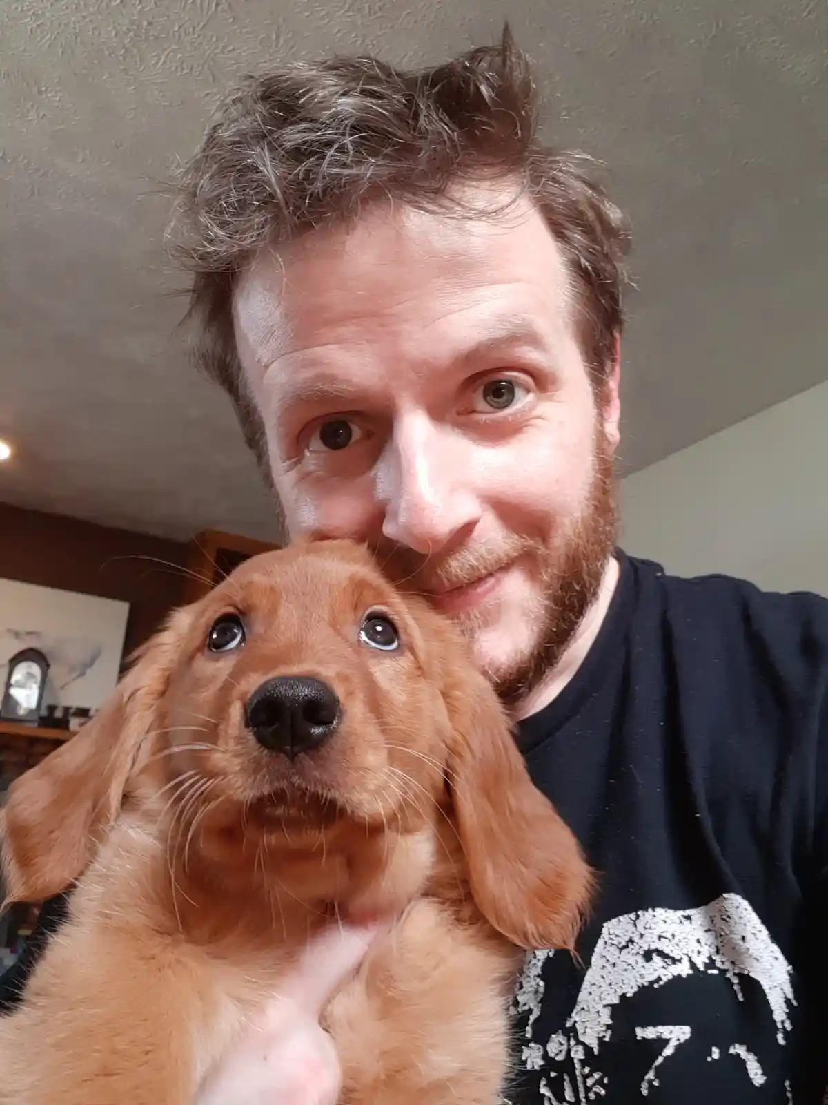 A man with a beard and messy hair holds a golden retriever puppy close to his face, both looking at the camera. The man has…