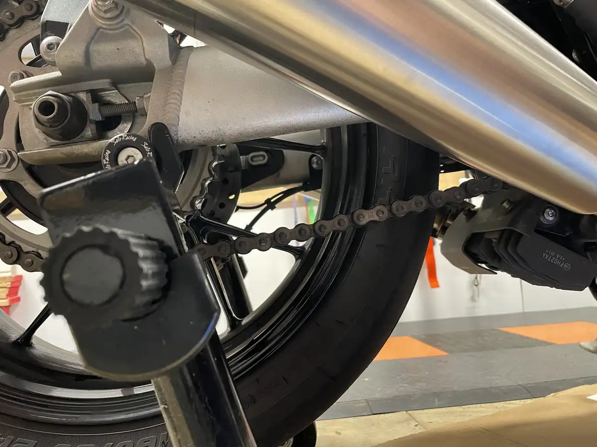 A close-up photo of a motorcycle's rear wheel, chain, and exhaust pipe, showing the chain resting on the tire. Overlaid text…