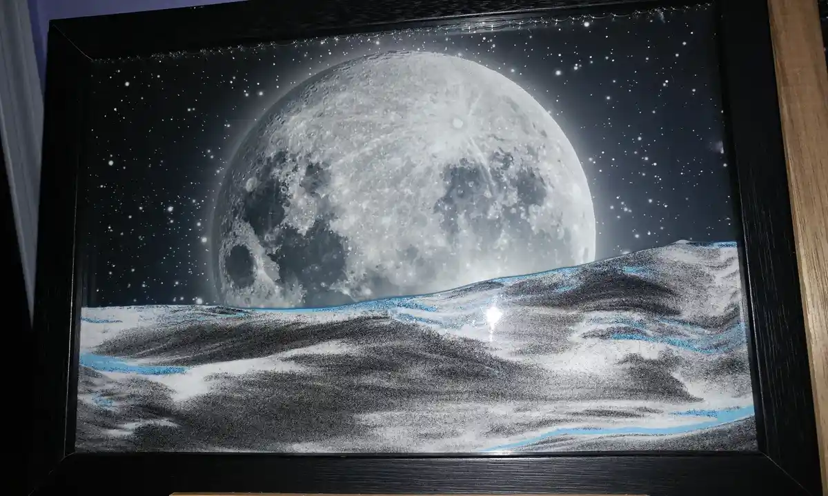 A framed artwork depicts a detailed, monochrome moon rising over a textured lunar landscape with blue glitter accents, set…