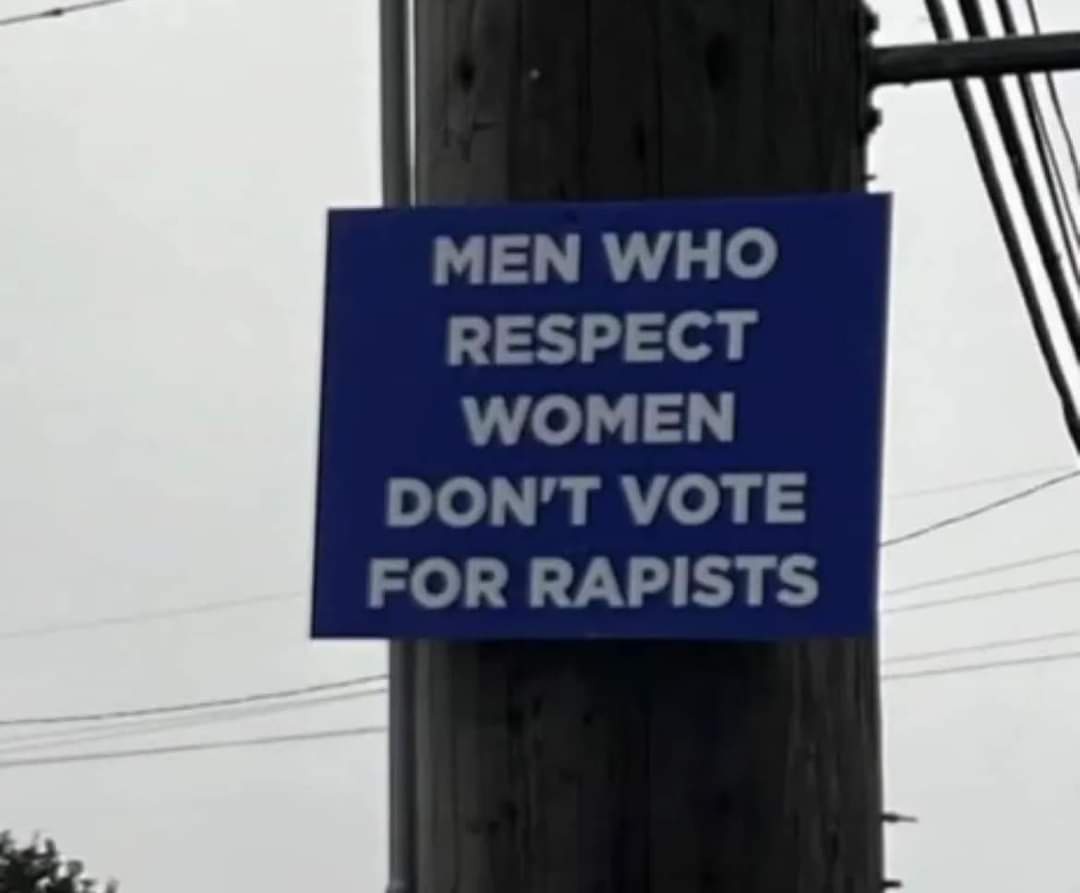 A blue rectangular sign with white text is taped to a wooden utility pole outdoors, surrounded by power lines and a gray…