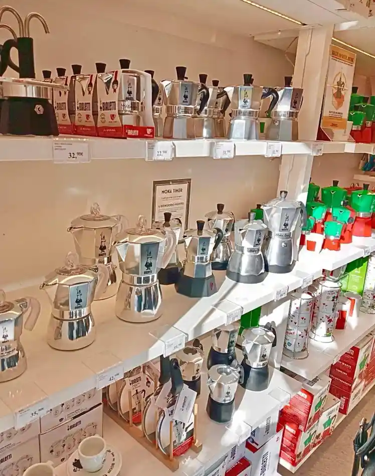 A store shelf displays numerous Bialetti moka pots in various sizes and colors, including classic silver, green, and red…