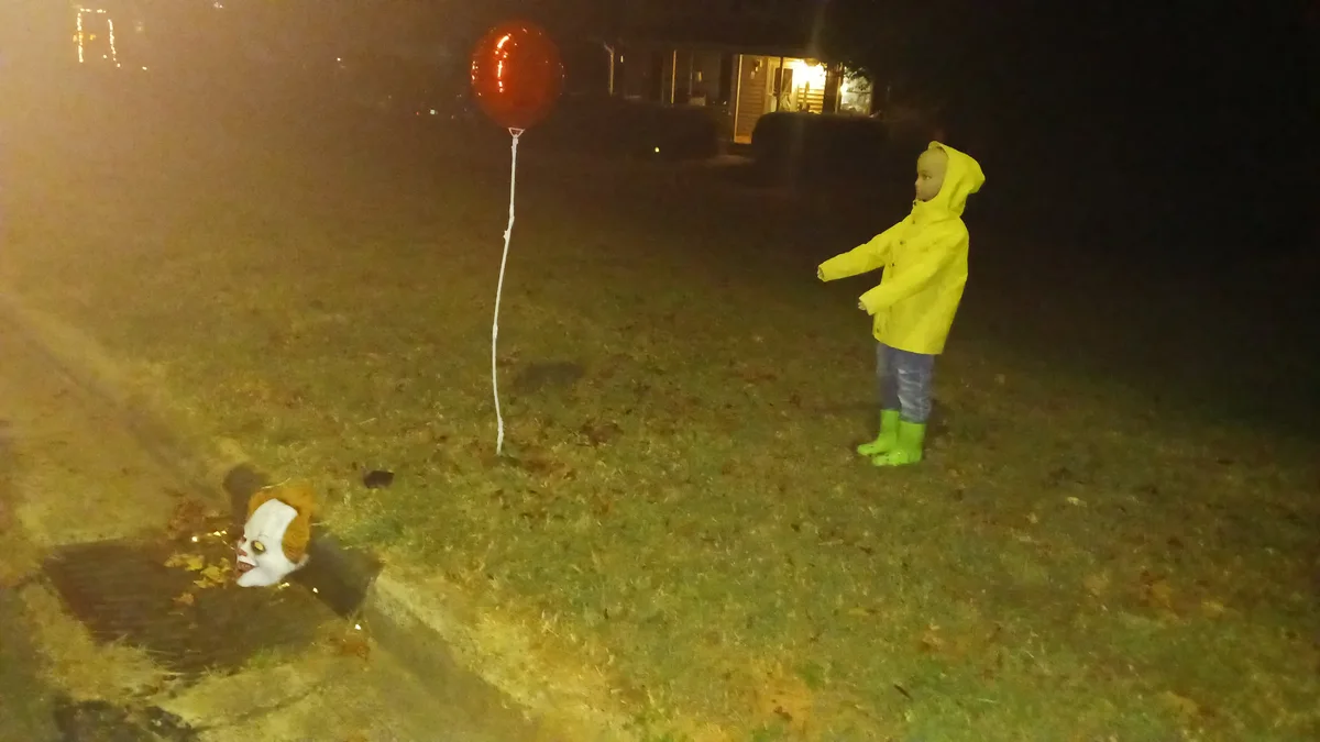 A child in a yellow raincoat and green boots stands on a grassy lawn at night, looking toward a red balloon on a white…