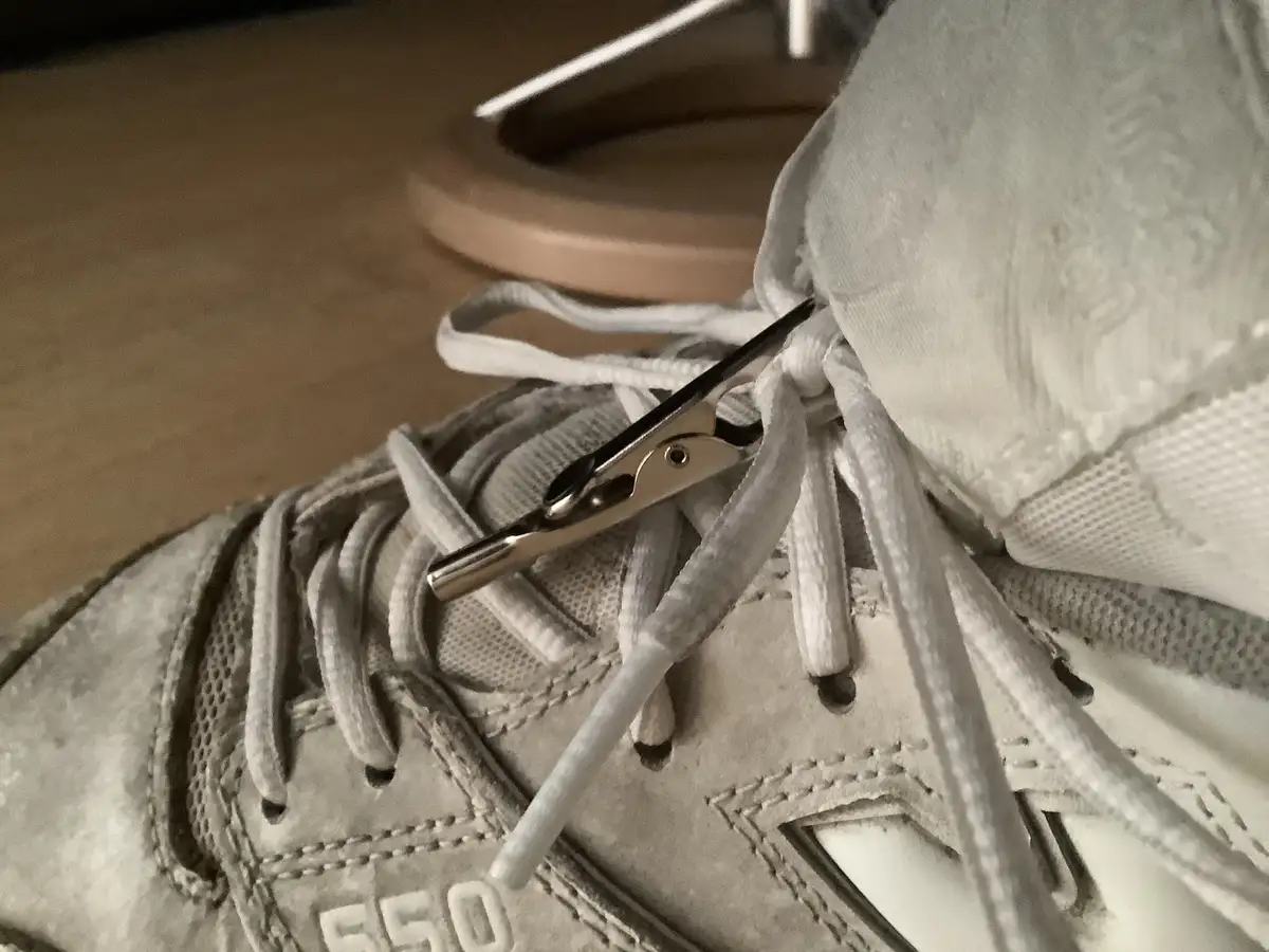 A close-up photo shows a silver metal clothespin clipped onto the laces of a white sneaker, holding the laces together at…