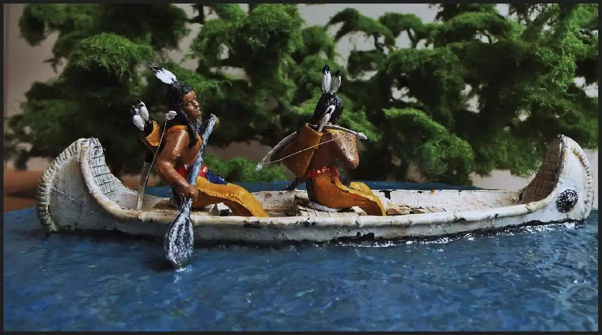 A diorama-style image shows two miniature figures in a canoe on a blue surface, with one figure resembling a Native American…