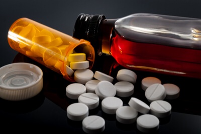An orange prescription pill bottle lies on its side with its cap off, spilling white pills onto a black surface. Beside it…