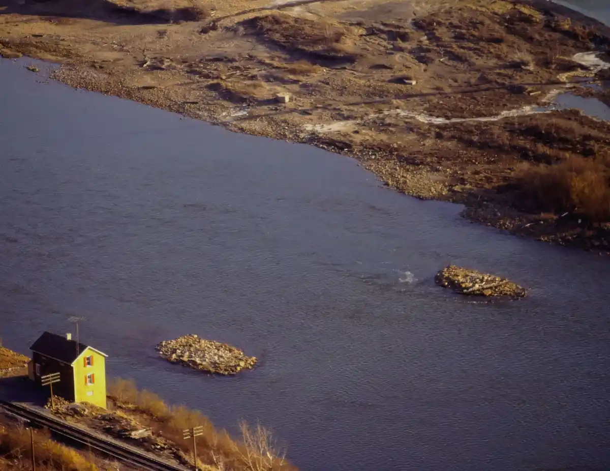 An aerial photo shows a small yellow house on the bank of a wide river, with two rocky islets in the water nearby. The…