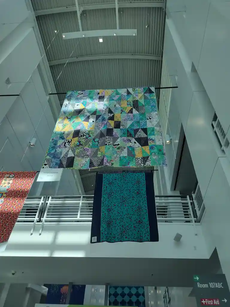 A colorful, geometric-patterned quilt hangs suspended from the ceiling of a modern, multi-level building atrium, with…