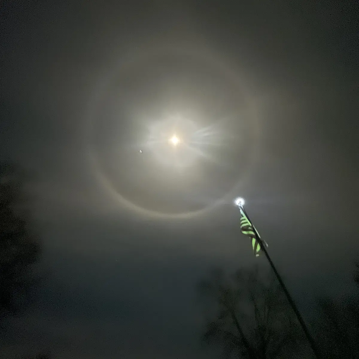 A nighttime photo shows a bright moon surrounded by a faint halo in a hazy sky, with a flagpole and American flag visible in…