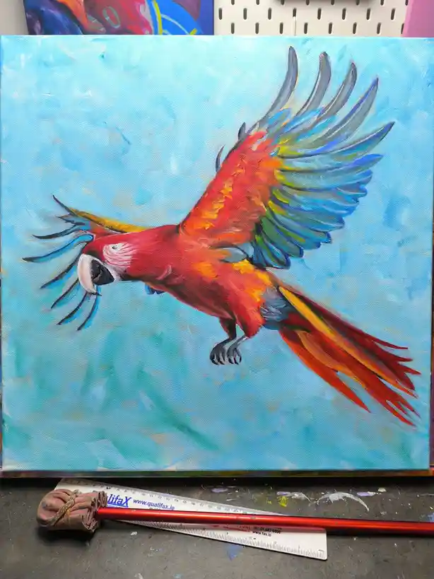 A vibrant oil painting of a red macaw with blue and green wings spread wide against a light blue sky, displayed on a canvas…