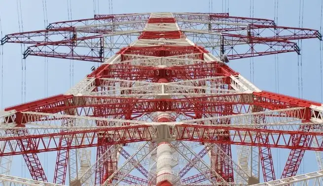 A low-angle photograph shows a massive, red-and-white lattice electricity pylon against a clear blue sky. The structure is…