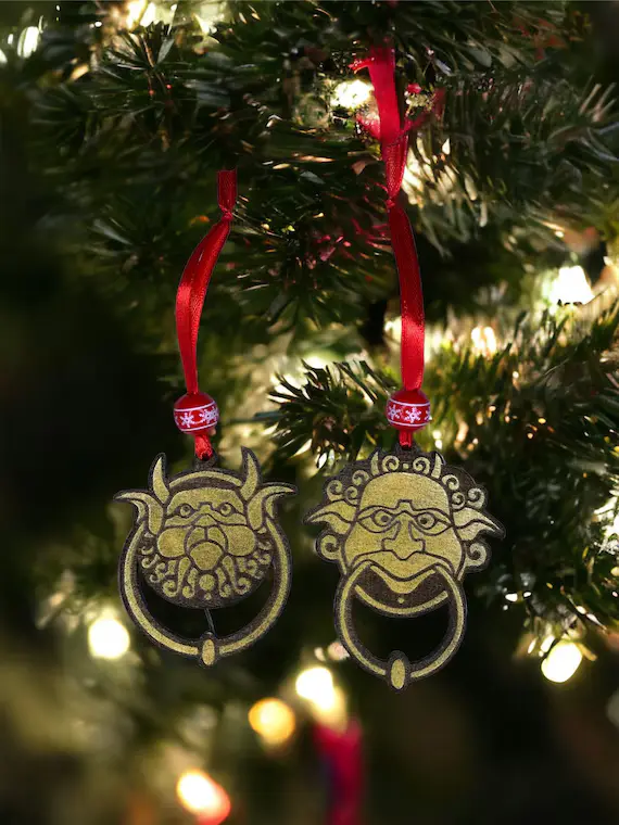 two ornate, gold-colored door knocker-shaped ornaments hang from red ribbons on a lit christmas tree, each depicting a…