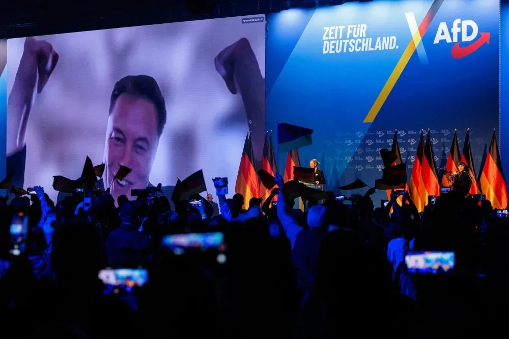 A large screen at a political rally displays a smiling Elon Musk with raised fists, while a crowd holds German flags and…