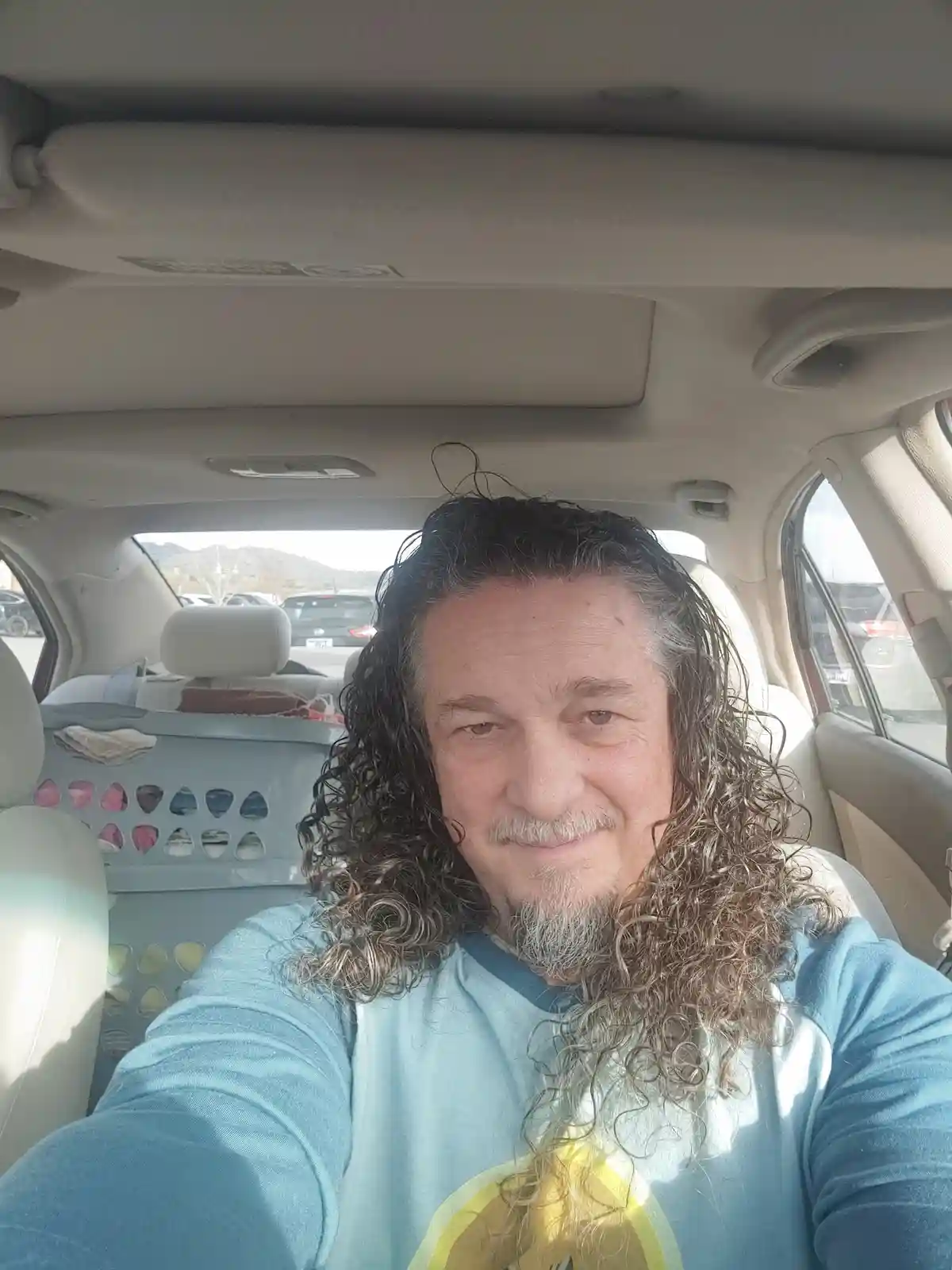 A man with long, curly, dark hair and a gray beard takes a selfie in the driver's seat of a car. Behind him, a light blue…