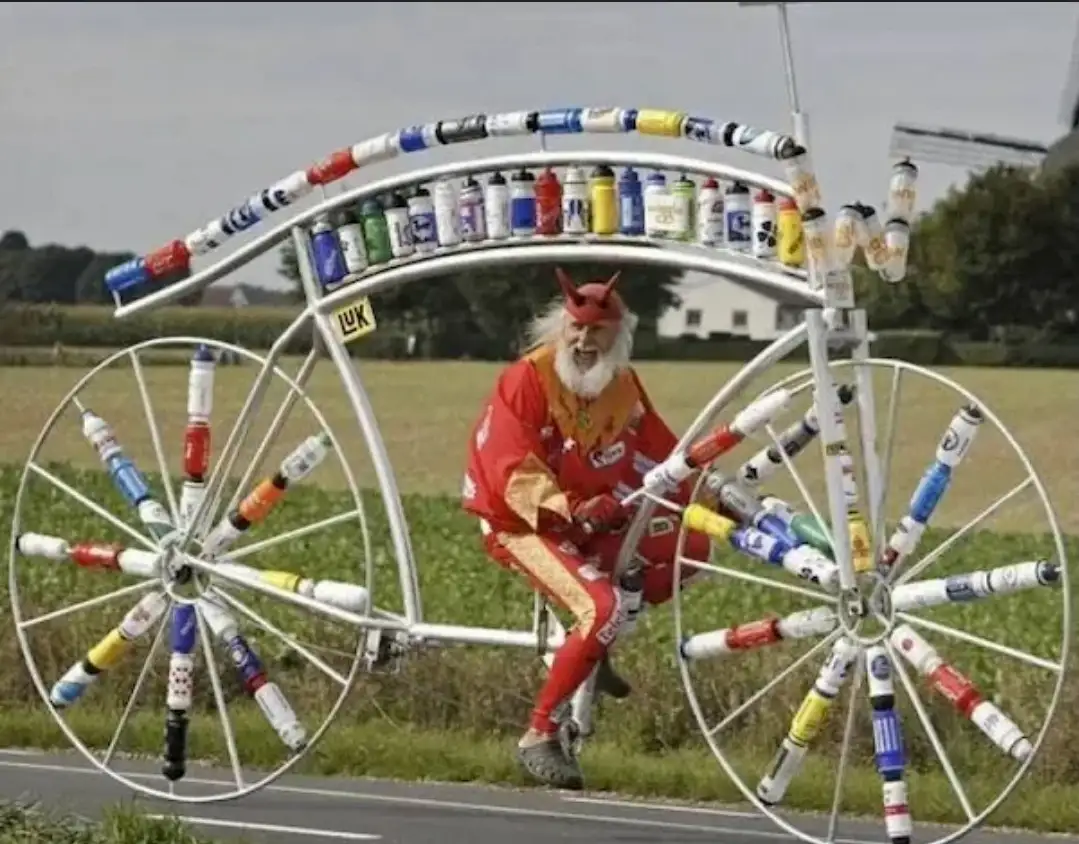 A man dressed in a red devil costume with a white beard and horns is riding a large, white, bicycle-like contraption. The…