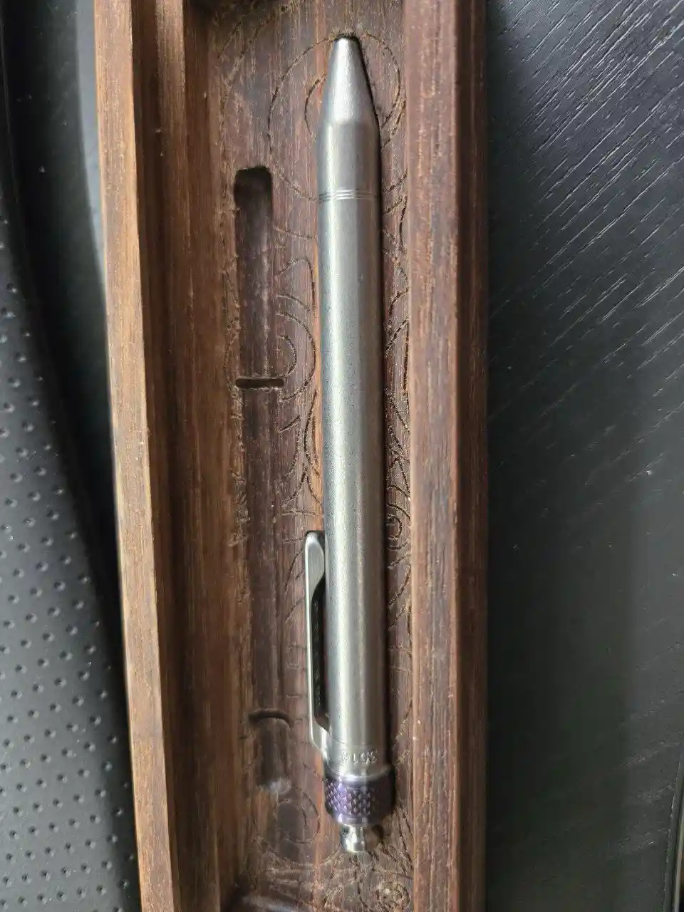 a silver-colored pen with a clip and textured grip is inserted vertically into a carved wooden holder, which is set against…