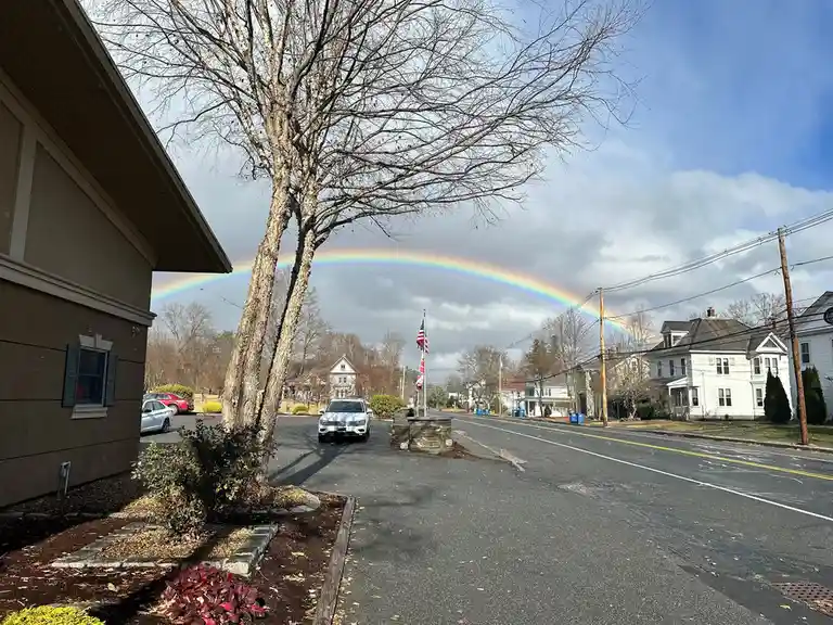 A residential street with houses, bare trees, and parked cars under a partly cloudy sky, featuring a vivid rainbow arcing…