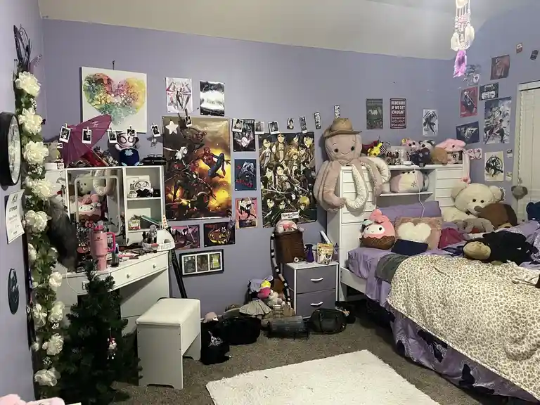 A cluttered bedroom with purple walls, covered in posters, plush toys, and decorations. A large octopus plushie wears a…