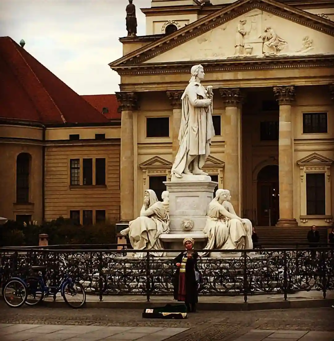 A person in traditional folk attire stands before a large classical statue in a public square, with a guitar case open on…