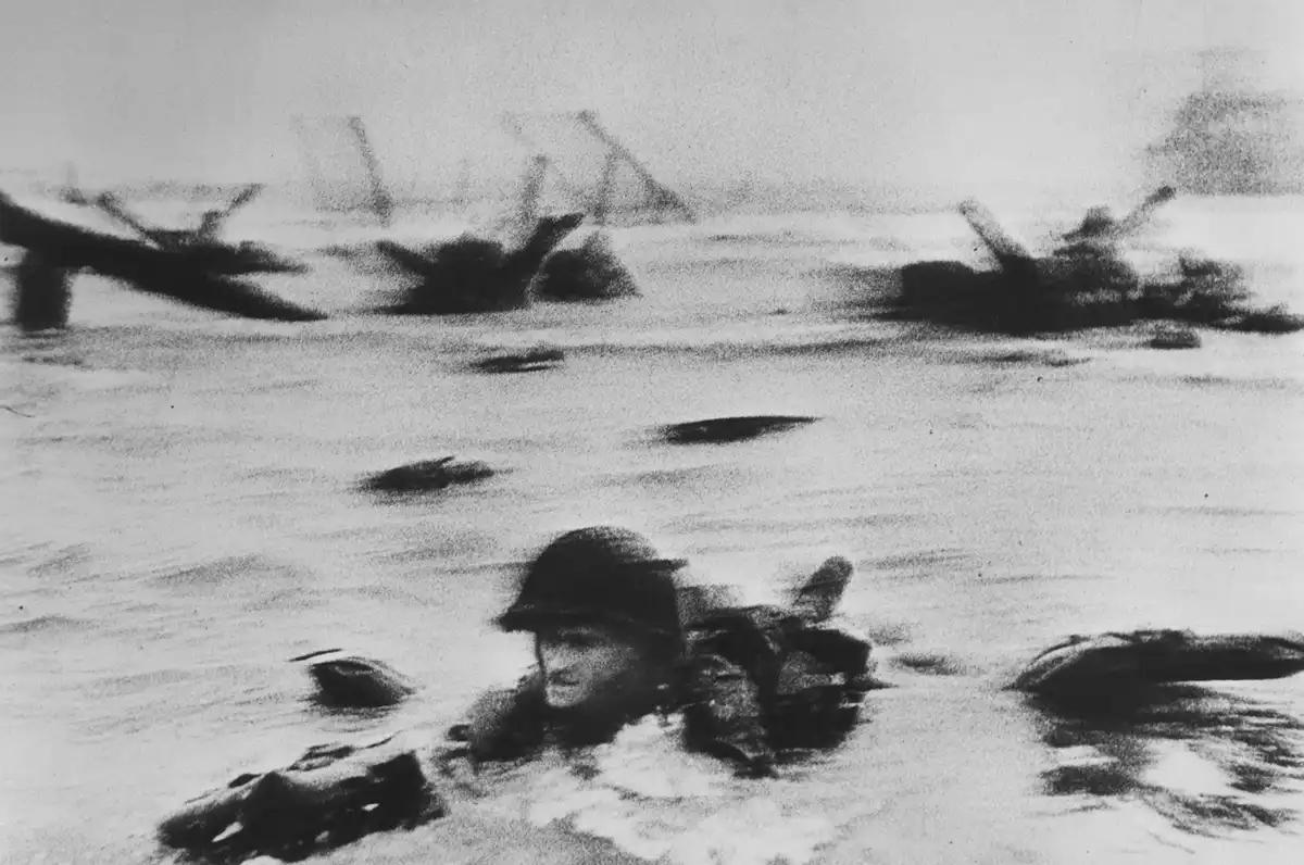 A grainy black-and-white historical photograph shows a soldier in a helmet wading through shallow water, surrounded by…